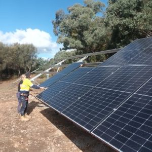 20240822_143112-scaled-qtauzk3jnv4j45cfps3ji39ryj0rzm36i39j0y9ifc Two trades attending to large solar panels on angle facing the sky, installed on the ground. Trees and sky in background.